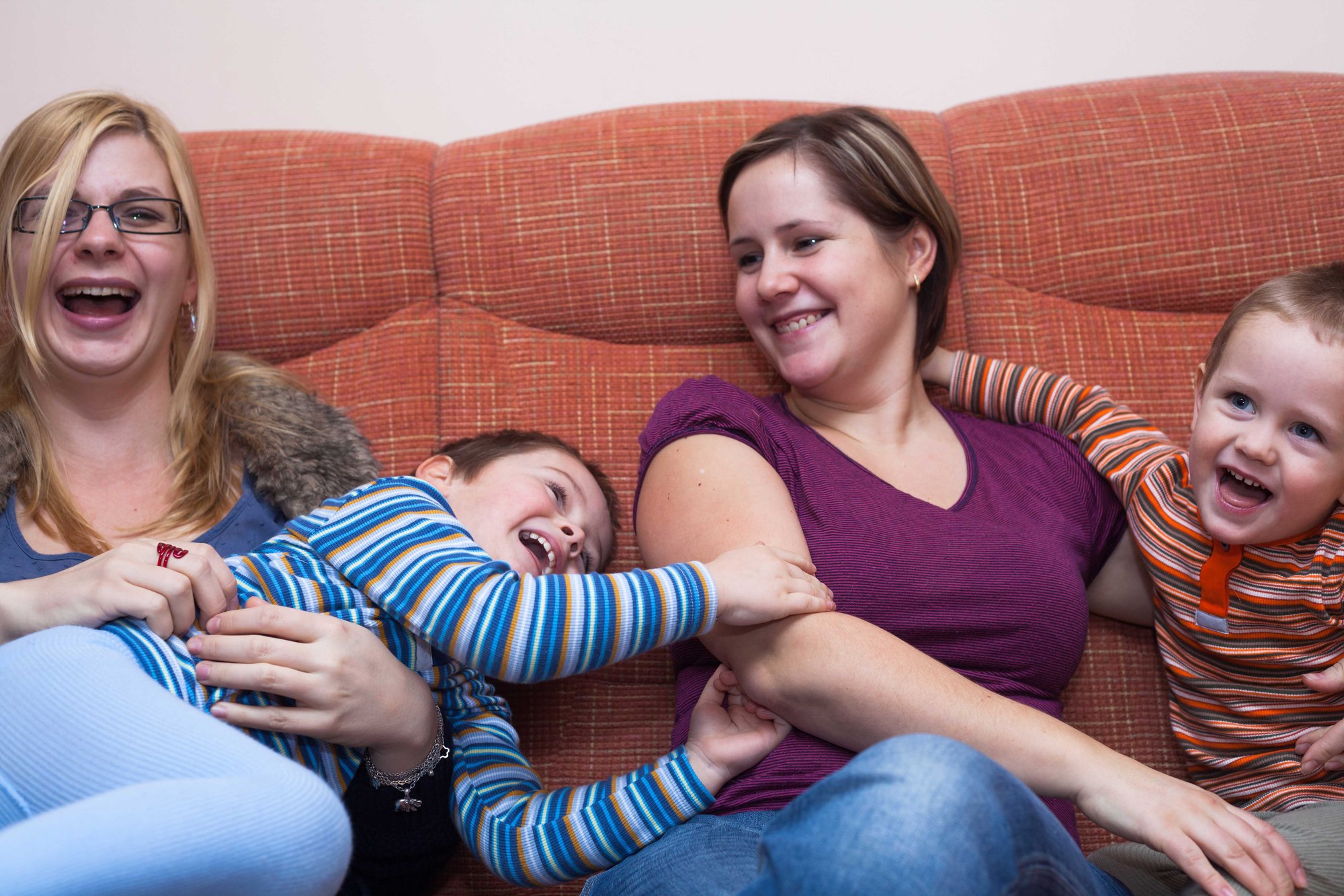 Two adult women and two children sitting on an orange couch smiling and laughing